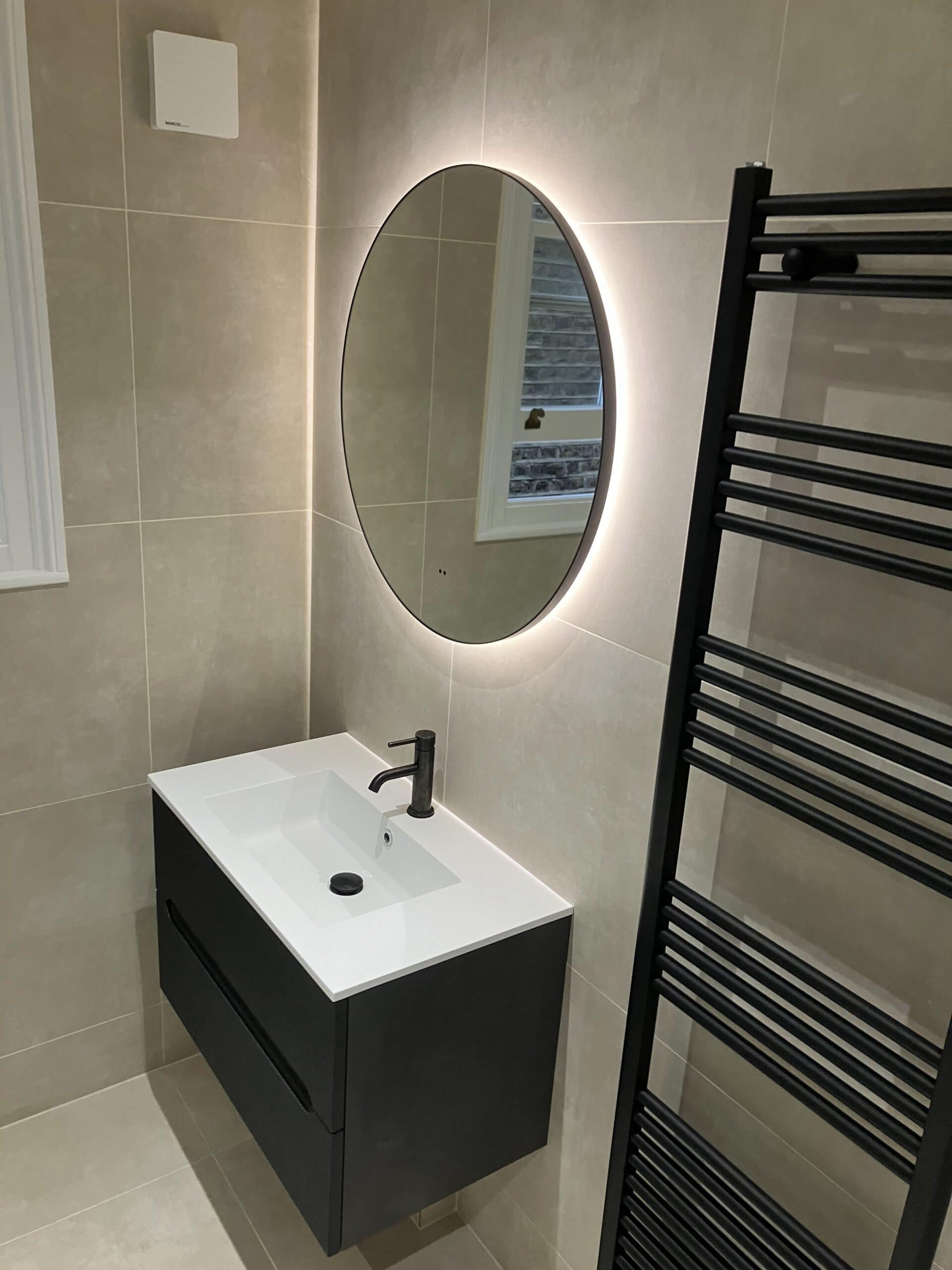 Wall-hung vanity unit with backlit oval mirror and black heated towel rail in a tiled bathroom